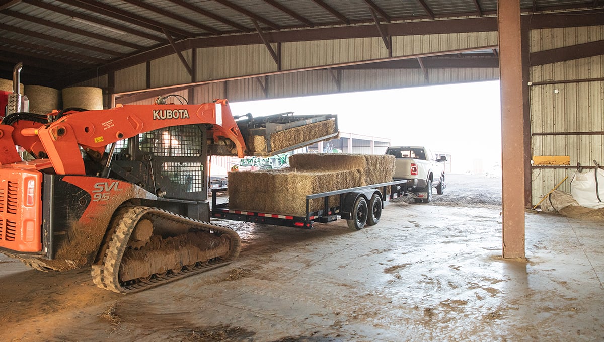 a kubota skid steer is loading hay into a 10pi tube top utility trailer