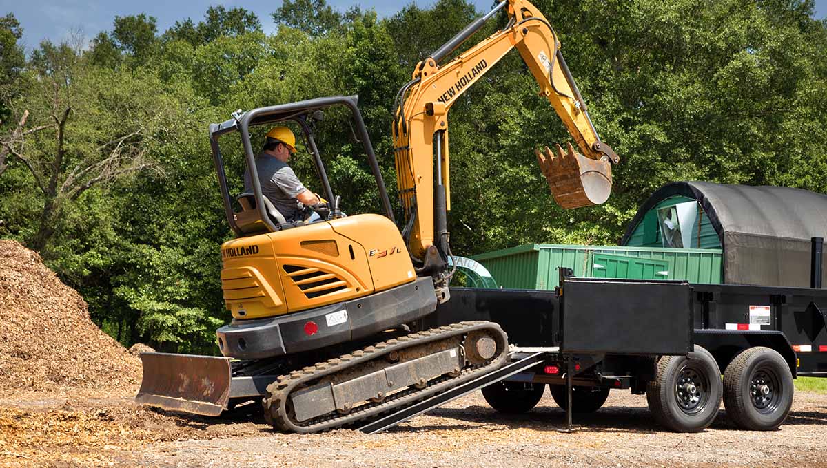 a man is driving a new holland excavator showcasing a 14TD commercial grade dump trailer