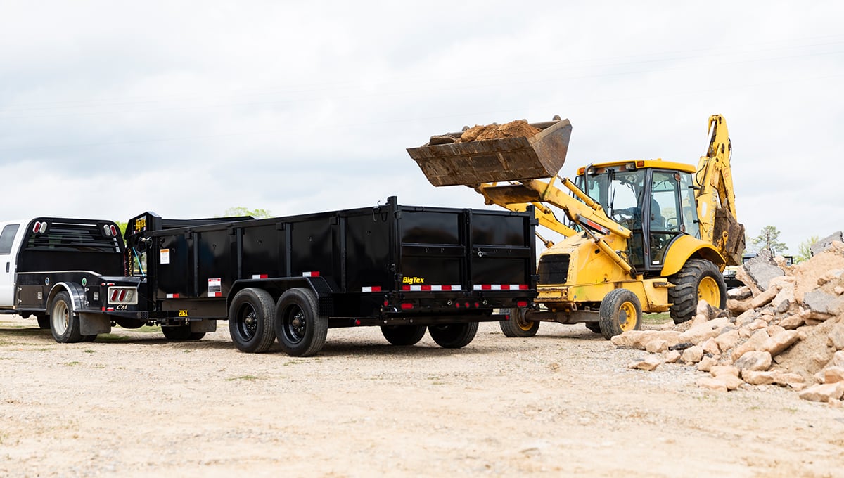 a yellow excavator is loading dirt into a 20gx commercial grade dump trailer
